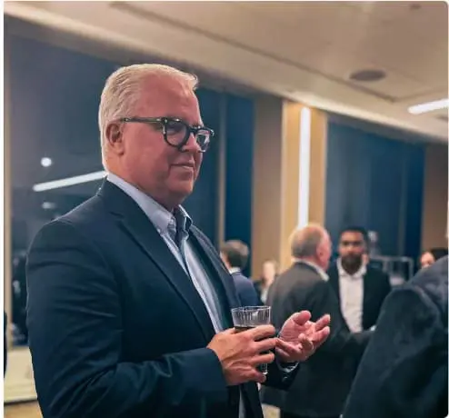 A man in glasses and a dark suit holding a glass and talking at a networking event featured on the home page.