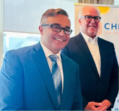 Two men in business attire smiling at a home page event, one in the foreground wearing a blue suit and glasses, and the other slightly blurred in the background.