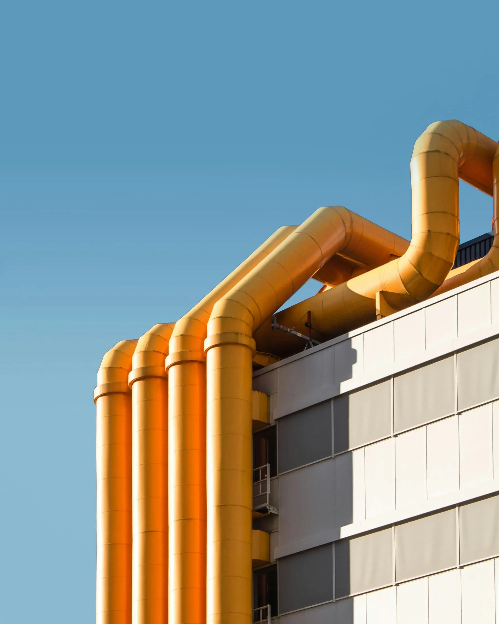 A building exterior with multiple large, yellow pipes extending from the roof down the side against a clear blue sky, part of an initiative by energy companies focused on decarbonizing.