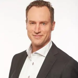 Bryce Conacher, dressed in a dark suit jacket and white dress shirt, poses against a plain white background, smiling slightly at the camera.