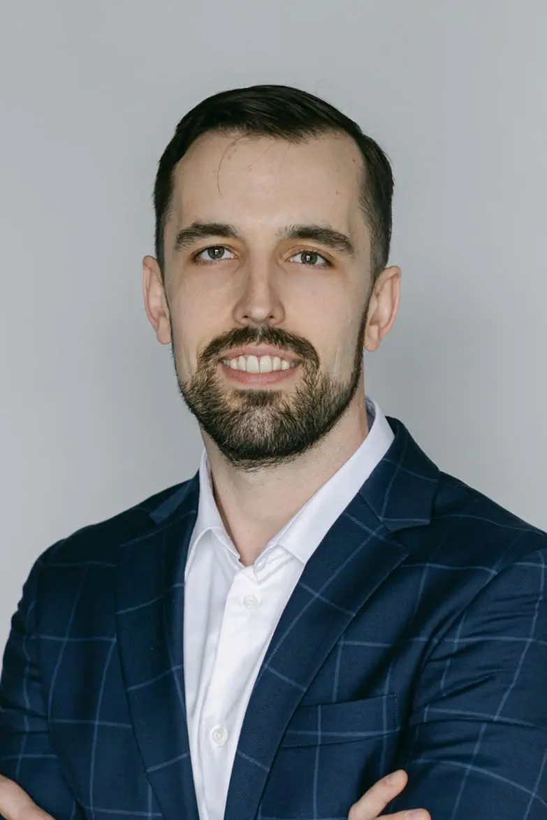 Matthew Small, a man with short dark hair and a beard, wearing a blue plaid suit jacket and white shirt, poses with arms crossed against a plain light background.