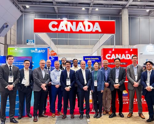 Group of professionals smiling on the home page in front of a booth labeled "Canada" at a trade show.