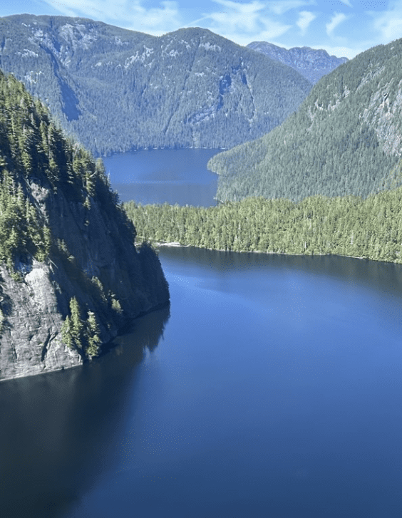 Aerial view of a large lake, part of the Inside Passage Marine, surrounded by dense forest and green mountains under a clear blue sky.