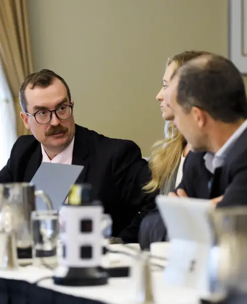 Three people in business attire sit at a conference table engaged in conversation about government relations, with documents, pitchers, and electronic devices in front of them.