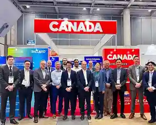 A group of people in business attire stand posing in front of a Canada booth at an indoor event or convention.