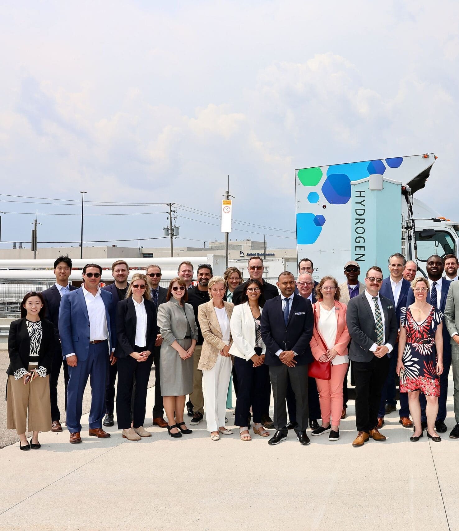 A large group of people in business attire stand outside in front of a hydrogen fuel sign and a truck, posing for a group photo on a sunny day.